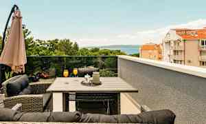 Terrasse med bord og stole, orange drikke i glas, parasol og udsigt over træer, bygninger og hav. (AI-genereret tekst)
