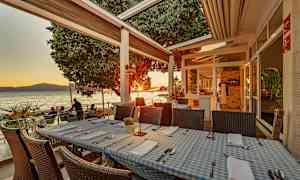 Restaurantborde med blå- og hvidternede duge på en terrasse ved havet under solnedgangen. (AI-genereret tekst)