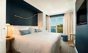 Et hotelværelse med en bred seng, hvidt sengetøj og en marineblå væg, store vinduer lukker naturligt lys ind og viser udsigt over tropisk vegetation og havet. (AI-genereret tekst)