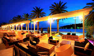 Udendørsmøbler på en terrasse med udsigt over havet og palmer om aftenen. (AI-genereret tekst)