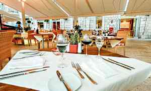 Et bord dækket til middagsgæster på en udendørsrestaurant med træmøbler og hvide tekstiler. (AI-genereret tekst)