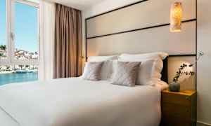 Hotelværelse med en dobbeltseng, pyntepuder og et natbord. Udsigt til vand og bygninger gennem vinduet. (AI-genereret tekst)