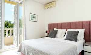 Et lyst soveværelse med en lyserød sengegavl, hvidt sengetæppe og en dør til en balkon. (AI-genereret tekst)