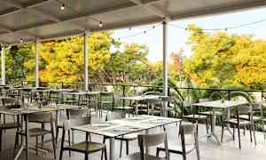 En udendørs restaurant med borde og stole under et tag, omgivet af grøntområde. (AI-genereret tekst)