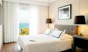 Et lyst hotelværelse med en dobbeltseng, moderne indretning og havudsigt gennem store vinduer. (AI-genereret tekst)