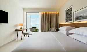 Hotelværelse med redt seng, skrivebord, stol og udsigt over havet. (AI-genereret tekst)