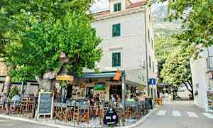 En udendørs café ved en bygning med hvide vægge og grønne skodder, omgivet af grønne områder og træer. (AI-genereret tekst)