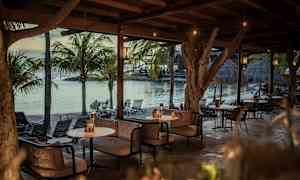 En udendørs lounge under et skur af træ med sofaer og borde ved havet, omgivet af palmetræer. (AI-genereret tekst)