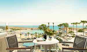 Udsigt fra en altan med bord og stole mod en strand med parasoller og palmer ved havet. (AI-genereret tekst)