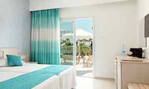 Lyst hotelværelse med dobbeltseng og udsigt til en terrasse med liggestole og parasoller. (AI-genereret tekst)