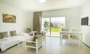 Lyst møbleret værelse med seng, stole, bord og udsigt til grønt område gennem store vinduer. (AI-genereret tekst)