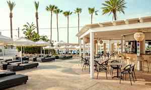 En udendørs servering med borde og stole under et hvidt tag, omgivet af solsenge og parasoller. I baggrunden ses palmer og blå himmel. (AI-genereret tekst)