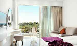 Et lyst hotelværelse med en sofa, en seng med lilla tæppe og en altan med udsigt over grønt område. (AI-genereret tekst)