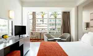 Hotelværelse med seng, siddepladser og balkon med udsigt over en gårdhave med palmer. (AI-genereret tekst)