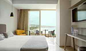 Et hotelværelse med moderne indretning, en bred seng, en gul stol, et bord med udsigt over stranden gennem store vinduer. (AI-genereret tekst)