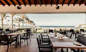 Restaurant med udendørsservering, palmer og hav i baggrunden. (AI-genereret tekst)