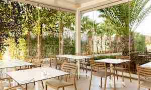 Billede af en udendørs servering med borde og stole omgivet af glasvægge og grønne omgivelser. (AI-genereret tekst)