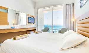 Hotelværelse med seng, skrivebord og balkon med udsigt over havet. (AI-genereret tekst)