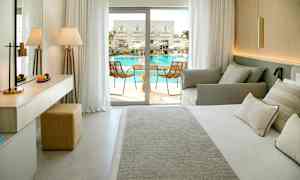 Et hotelværelse med seng, sofa og adgang til en balkon med udsigt til poolområdet. (AI-genereret tekst)