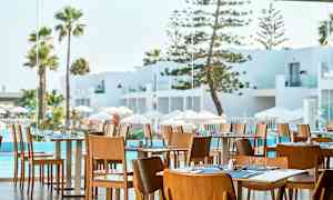 Restaurant med træstole og dækkede borde, udsigt til hvid arkitektur, pool og palmer. (AI-genereret tekst)