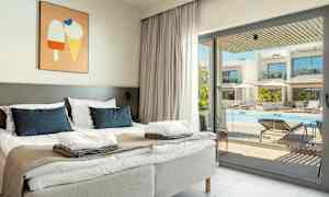 Et hotelværelse med en dobbeltseng, lysegrå sengegavle og blå puder. Et billede med ismotiv hænger på væggen. Gennem glasdøren ses en udendørs pool med liggestole og parasoller. (AI-genereret tekst)