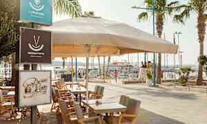 Udendørs servering med borde og stole under en stor parasol ved en havn. (AI-genereret tekst)