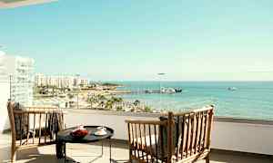 To træstole og et bord på en hotelaltan med udsigt over strand og hav. (AI-genereret tekst)