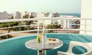 En balkon med udsigt over by og hav. Et bord med et glas, et grønt æble, en bog og solbriller. (AI-genereret tekst)