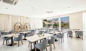 Et lyst og moderne restaurantområde med runde og firkantede borde omgivet af polstrede stole. Store vinduer viser en udendørs servering. (AI-genereret tekst)