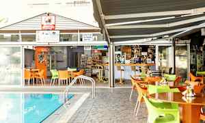 Udendørsområde med borde og stole ved en pool, med en bar og spisesteder i baggrunden. (AI-genereret tekst)