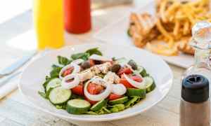 En tallerken med salat indeholdende tomat, agurk, løg, fetaost og oliven. I baggrunden ses pommes frites og sandwich. (AI-genereret tekst)