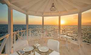 En udendørsrestaurant med hvide møbler, beliggende ved havet, med solnedgang i baggrunden. (AI-genereret tekst)