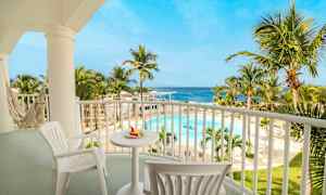 Billedet viser en afslappende altan med bord og stole samt en hængekøje. Udsigten er over en tropisk pool omgivet af palmer og havet i baggrunden. (AI-genereret tekst)