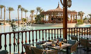 Et dækket bord på en veranda med udsigt over en pool omgivet af palmer og en pavillon. (AI-genereret tekst)