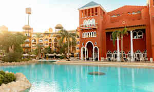 Hotelbygning med rød facade ved et poolområde med liggestole og parasoller. (AI-genereret tekst)