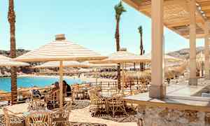 Udendørsservering med bambusstole og parasoller nær stranden med blåt vand. (AI-genereret tekst)