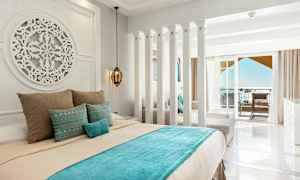 Et hotelværelse med lyse vægge, en dekoreret seng med blå og beige sengetøj, samt en altan med havudsigt. (AI-genereret tekst)