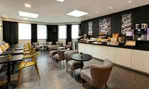 Et caféområde med borde, stole og en bænk langs væggen. Et serveringsbord med morgenmadstilbehør og en kaffemaskine ses i baggrunden. (AI-genereret tekst)