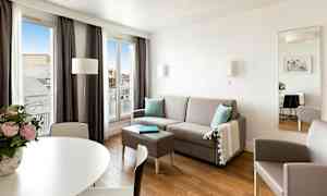 En moderne, lys stue med grå sofa, bord, lænestol og store vinduer, der lader naturligt lys komme ind. (AI-genereret tekst)