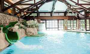 En indendørs pool med en vandrutsjebane nær en klippe og et kunstigt vandfald. Poolområdet er omgivet af store vinduer og trækonstruktioner. (AI-genereret tekst)