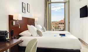 Et hotelværelse med dobbeltseng, skrivebord og en åben balkondør med udsigt over de omkringliggende bygninger. (AI-genereret tekst)