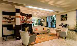 Et opholdsområde med bløde lænestole, bord og sofa på et orange gulvtæppe, med planter og kunst på væggene. Stor spejl og vinduer. (AI-genereret tekst)