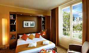 Et hotelværelse med en dobbeltseng, puder og tæpper i varme farver samt en stor vinduesudsigt til træer og bygninger. (AI-genereret tekst)