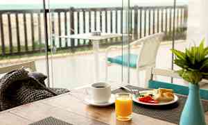 Morgenmadsbord med kaffe, juice og grøntsager nær et glasvindue med udsigt over en terrasse med havet i baggrunden. (AI-genereret tekst)