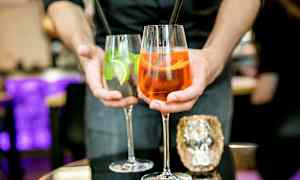 To drinks holdes frem af en person, et glas indeholder en orange cocktail og det andet en klar cocktail med lime og mynte. (AI-genereret tekst)
