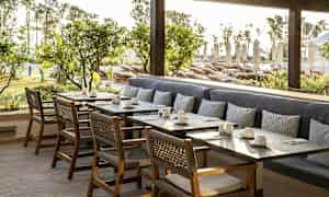 Udendørsområde med borde, stole og bænk med puder foran grønne områder og solvogne. (AI-genereret tekst)