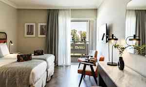 Hotelværelse med to enkeltsenge, siddeområde og skrivebord. Balkon med udsigt over træer. (AI-genereret tekst)