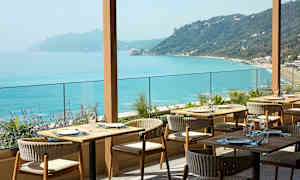 En restaurant med udendørs servering og udsigt over turkisblåt hav og kuperet kystlinje. (AI-genereret tekst)