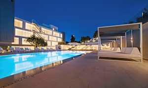 En udendørs pool med liggestole og moderne siddepladser, omgivet af et oplyst hotel under nattehimlen. (AI-genereret tekst)