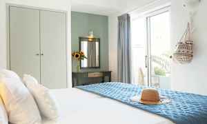 Et lyst hotelværelse med en redt seng, en solhat på blåt sengetæppe og et dekorativt bord med spejl og en vase med solsikker. (AI-genereret tekst)